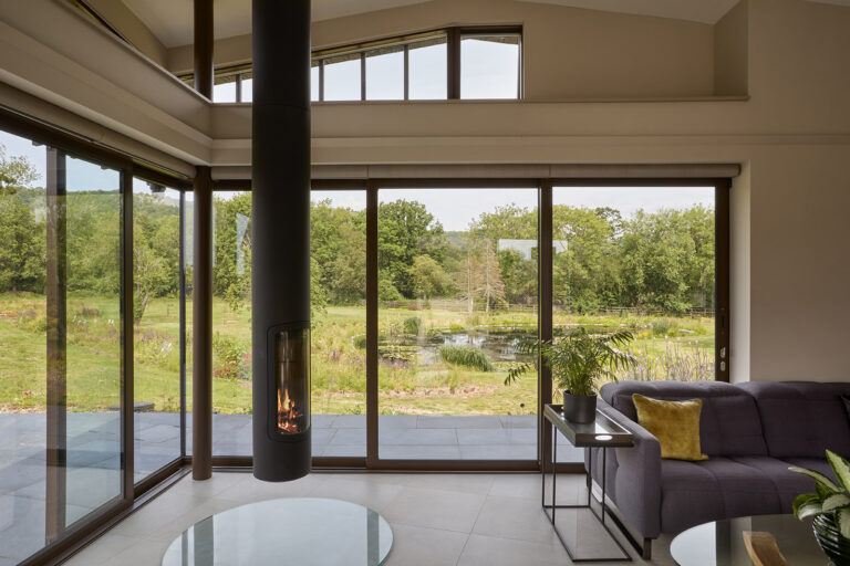 Living room at bespoke build house Ridgemere looking out through tall glass windows and doors onto the surrounding landscape.